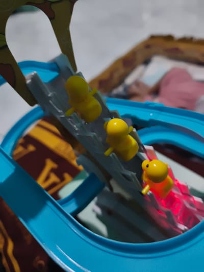 The image shows a close-up, elevated view of a yellow duck toy race track. Three yellow duck figurines are positioned on the track. The duck at the front is illuminated by a red light from below. The track itself is blue and gray, with a yellow and brown patterned object visible underneath it. A person is partially visible in the background, wearing a pink garment. The scene appears to be indoors, possibly a home environment, and there is no indication of the specific location within Barengkrajan, Indonesia.