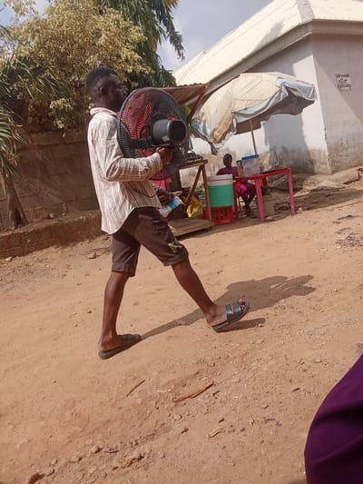 A man walks across a dusty, dirt path in Kagini, Nigeria, carrying a large black and red fan. He is wearing a vertically striped, long-sleeved shirt and brown shorts, with black sandals on his feet. The sun is bright, casting a shadow of the man and his fan onto the ground. In the background, a woman sits at a red table under a tan and blue umbrella, with a cooler and drinks visible. Behind her, a building with a light-colored roof stands. The scene appears to be outdoors, possibly a market or roadside stall, during the daytime. A sign on the building reads "SELF CONTAIN 4 RENT" followed by a phone number.