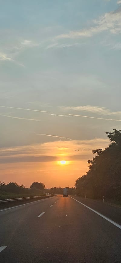 The image captures a sunset over a highway in Balatonszabadi, Hungary. The sun, a bright orange orb, is low in the sky, partially obscured by wispy clouds. The sky transitions from a pale blue at the top to shades of orange and yellow near the horizon, reflecting the warm glow of the setting sun. Several white contrails are visible in the upper portion of the sky.

The highway stretches forward, with a single lane visible in the foreground, marked by white lines. The road surface is asphalt, appearing dark and wet in places, reflecting the sunset's light. On the right side of the road, a dense line of trees and bushes creates a dark silhouette against the vibrant sky. On the left side, a guardrail separates the highway from a grassy embankment, beyond which more trees are visible.

A white truck with a cylindrical tank is traveling away from the viewer on the highway. It is illuminated by the setting sun, casting a long shadow on the road. Other vehicles are faintly visible in the distance on the highway. The overall scene depicts a serene, yet dynamic, moment on a rural highway at dusk.