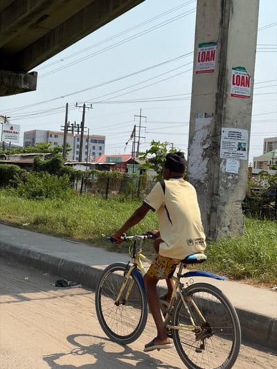 The image depicts an outdoor daytime scene in Ikota, Nigeria, under a concrete overpass. A young male, visible from the back and right side, rides a light yellow bicycle on an unpaved dirt shoulder alongside a concrete curb. He wears a black beanie, a light yellow short-sleeved t-shirt with blue accents and a UNHCR logo on the back, patterned shorts, and dark slides. The bicycle has a yellow frame, blue handlebars, a blue seat, and a blue rear fender, appearing worn.
To the rider's left, a strip of overgrown green grass and weeds separates the shoulder from a fence covered in foliage and beyond that, low-lying structures with corrugated roofs. In the background, taller multi-story buildings with blue-tinted windows are visible. Numerous utility poles and a dense network of overhead power lines crisscross the clear, light blue sky. A white "Dillon Gas" billboard is partially visible on the far left.
To the rider's right, a large, light-colored concrete pillar stands prominently, adorned with multiple posters. Two prominent red and white posters advertise "Get an Instant LOAN," while another poster offers "AFFORDABLE LAND & HOMES." Strong shadows are cast on the dirt surface by the rider and bicycle, indicating bright sunlight.