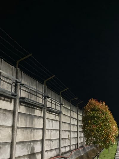 The image depicts a close-up view of a rooftop at night, with a dark, featureless sky dominating the left side of the frame. On the right, a section of a tiled roof is visible, laid out in a grid pattern. Several metal railings or safety barriers are present on the roof, extending upwards and to the right, casting shadows on the tiled surface. A lush green and reddish-brown bush is situated on the far right of the rooftop, its foliage illuminated by an unseen light source. A red cable or hose runs along the edge of the roof. The scene appears to be outdoors, possibly in Gambut Barat, Indonesia, given the context. The time of day is clearly night due to the deep blackness of the sky. There are no people or animals visible in the image. The overall impression is one of an industrial or functional rooftop area at rest, illuminated by artificial light.