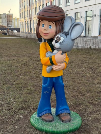 A full-length, outdoor photograph depicts a brightly colored statue of a boy holding a rabbit in Yanino-1, Russia. The central statue portrays a cartoonish young boy with brown hair, blue eyes, and a slight smile, clad in a yellow long-sleeved shirt with green accents, blue trousers exhibiting dirt marks, and brown shoes. He cradles a grey cartoon rabbit, characterized by large ears, brown eyes, a white snout, and a black cross on its forehead. The statue rests on a green, textured circular base. The immediate foreground features sparsely vegetated, exposed soil. In the background, modern multi-story residential or commercial buildings with light facades are visible. A construction site, complete with an unfinished building frame and a tower crane, occupies the far left background. Multiple parked vehicles are present in the mid-ground. The scene is illuminated by ambient daylight.