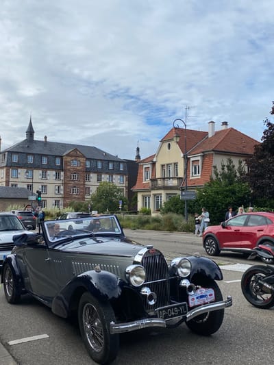The image depicts a street scene in Molsheim, France, during the "XIII Festival Bugatti."

In the foreground, a two-tone vintage Bugatti convertible, featuring a dark grey body with dark blue fenders and running boards, is positioned on an asphalt road. The vehicle has chrome headlights, a chrome grille with the "Bugatti" emblem, and a chrome front bumper with "CHOPARD" embossed on the left. A French license plate "AP-049-YL" is affixed to the front, above which a small event plate reads "XIII FESTIVAL BUGATTI." Two individuals occupy the front seats; the driver, an older male, wears a light-colored hat, and the passenger wears a dark face covering. To the immediate left, the front of a silver modern car is partially visible. To the right, the front wheel and engine section of a dark motorcycle are visible.

In the midground, a modern red SUV is parked on the side of the road. Several bystanders observe the scene from a paved area to the right, including at least two adults and a child. Further down the road, additional vehicles are visible in traffic.

The background features characteristic European architecture. On the left, a large, multi-story building with a dark slate-tiled hip roof and a central square tower with a pointed spire dominates the skyline. Its facade is a mix of light-colored and reddish-brown sections with numerous windows, many fitted with light shutters. On the right, a two-story house with a distinctive reddish-brown tiled mansard roof, dormer windows, and a light yellow stucco facade, including a second-story balcony, is present. A curved street lamp pole extends over the road near this house. Another distant spire is visible beyond the central background. Green trees and shrubs line the street between the road and buildings. A green traffic light is visible on the left. Road signs, including one indicating "Molsheim," are present. The sky is overcast with scattered clouds.