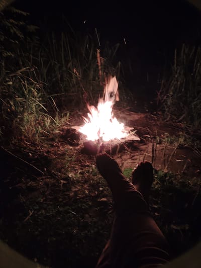 The image depicts a nighttime scene with a campfire burning in the center. Tall, dark grass and foliage surround the campfire. In the foreground, two bare feet and lower legs are visible, suggesting a person is sitting by the fire. The lighting is dim, with the fire providing the primary light source. The environment appears to be outdoors, likely in a natural or rural setting. There is no specific indication of the city of Balyko-Shchuchynka, Ukraine.