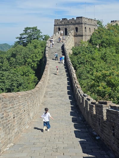 The image depicts a section of the Great Wall of China, likely located in the Huairou District of China, under a bright, partly cloudy sky. The stone structure of the wall, with its characteristic crenellations and watchtowers, snakes across a lush, green, mountainous landscape.
In the foreground, a young girl with braided hair, wearing a white t-shirt and blue pants, runs away from the camera along the paved walkway of the wall. Her shadow is cast on the stones behind her. Further up the wall, several other individuals, appearing to be tourists, are walking in both directions. Some are seen in groups, while others are walking individually. They are dressed in casual clothing suitable for warm weather.
The wall itself is constructed of dark grey bricks, with some sections showing signs of wear and tear. Watchtowers, some with arched windows, punctuate the length of the wall, indicating its historical defensive purpose. Dense green foliage, composed of trees and shrubs, surrounds the wall, suggesting it is a well-preserved and accessible tourist site. The path is made of large, flat stone slabs, which are slightly uneven and show cracks in places. The scene conveys a sense of historical grandeur combined with modern tourism.
