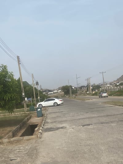 The image shows a street scene in Iddo Tudunwada, Nigeria. The view is from an elevated position, possibly on a bridge or elevated walkway. A white car is parked on the side of the road next to a green trash can. Power lines and utility poles run along the side of the road. In the distance, there are buildings and a hill or mountain. The sky is overcast. The road appears to be asphalt and is showing some signs of wear. There is a concrete wall or structure in the foreground.