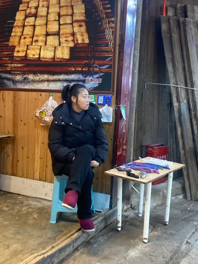 A woman rests on a blue chair outdoors. She is wearing a black jacket, black pants, and maroon slippers. Behind her is a food advertisement and a plastic bag containing food. To her side is a table displaying various items, with a red fire extinguisher nearby. The setting appears to be an outdoor market or street food area.