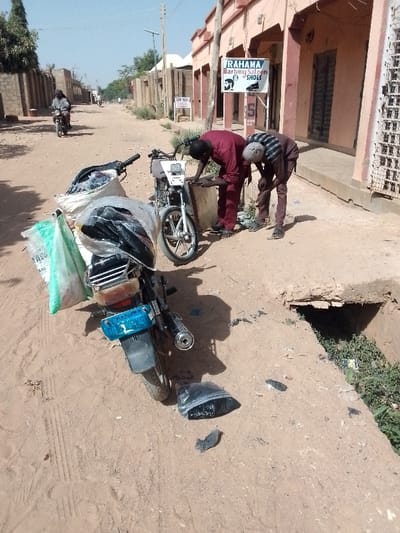 The image captures a sunny, mid-day scene on a dusty, unpaved road in what appears to be a town or village. On the right side of the road, there's a light reddish-brown building with a flat roof, which houses a business. A prominent sign on this building reads "RAHANA Barbing Saloon & SHOES," accompanied by icons of a man's head (for barbing) and a shoe (for shoes).
In the foreground, two men are bent over, appearing to inspect or work on a light-colored motorcycle parked beside the road. One man, wearing a maroon long-sleeved shirt and pants, is leaning over the front of the motorcycle. The other man, dressed in a striped long-sleeved shirt and dark pants, is similarly bent over, looking down near the motorcycle's engine area. Both men are wearing sandals.
To the left of these men, another black motorcycle is heavily loaded with large white and clear plastic bags filled with goods, possibly merchandise, suggesting it's used for transport or delivery. The bags are stacked high on the seat and rear rack. The rear license plate of this motorcycle is blue with white characters, which appear to partially read "FCT ABJ," indicating Abuja, Nigeria.
Adjacent to the building on the right, there's a visible concrete drainage ditch or open sewer with some green vegetation growing within it. The ground is dry and dusty, with visible tire tracks. Scattered debris, including pieces of dark plastic, are on the ground near the motorcycles and the ditch.
In the background, further down the road, another person is seen riding a motorcycle away from the viewer. On the left side of the road, there are walls or fences made of concrete blocks, and a utility pole stands tall. The clear sky and strong shadows indicate bright, direct sunlight.