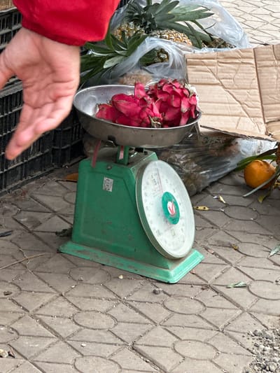 A person's hand wearing a red sleeve is visible in the upper left of the frame, reaching towards the left and out of frame. The hand is open and appears to be in motion. The focal point is a green mechanical scale placed on a patterned tiled ground. A metal bowl sits atop the scale, filled with bright pink dragon fruits. The scale has a circular dial with markings and a needle indicating a weight. In the background, several pineapples are visible, some enclosed in clear plastic bags. A single orange fruit rests on the ground to the right of the scale. Cardboard boxes are also present in the background. The scene depicts a market stall or roadside produce stand, likely in Bắc Yên, Vietnam, given the context provided. The lighting suggests it is daytime.
