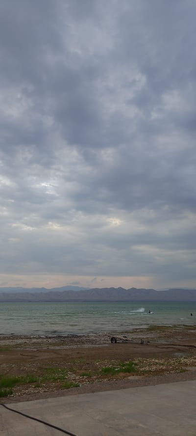 The media file shows a body of water, likely a lake or sea, with a distant mountainous landscape. The sky is overcast with dramatic, grey clouds, suggesting a possibly stormy or late afternoon/early morning atmosphere. On the water, a person is visible on a jet ski, creating a spray as they move. The shoreline is a mix of sand, pebbles, and some sparse green vegetation, with a paved area to the right of the frame. A trailer with wheels and some wooden planks is parked on the shore. The overall scene evokes a sense of a natural landscape with recreational activity. The location context suggests Chigdalik, Tajikistan.