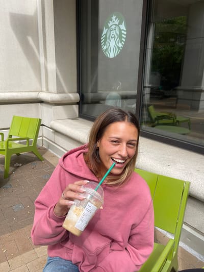 A young adult female is depicted seated outdoors on a lime-green metal chair, on a paved surface next to a building. She is wearing a pink hooded sweatshirt and blue jeans. Her light brown hair, with visible lighter streaks, frames her smiling face as she looks directly at the camera. She holds a clear plastic Starbucks cup, containing a partially consumed iced beverage and a green straw, to her mouth. The cup features a white label with "Lira" handwritten on it and a small golden star decal. A large green Starbucks siren logo is prominently displayed on the glass window of the building behind her, reflecting outdoor elements. The building exhibits light-colored stone or concrete architectural details. This scene is situated in Arlington, United States.