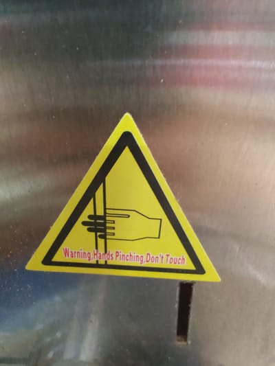 The media file is a close-up, angled shot of a yellow triangular warning sticker with a black border and black graphic. The sticker is affixed to a brushed metal surface. The graphic on the sticker depicts a stylized hand with fingers being crushed between two horizontal lines, symbolizing a pinching hazard. Below the graphic, in red, is the text "Warning, Hands Pinching, Don't Touch." The metal surface has visible vertical brush strokes and some faint light reflections, suggesting it might be part of a machine or appliance. To the right of the sticker, there is a narrow, dark horizontal slit in the metal surface. The lighting appears to be artificial, and the overall setting is likely industrial or a workshop, given the presence of warning labels and machinery elements. The image does not contain any people or specific environmental cues for time of day or weather. The scene is static, focused solely on the warning sticker and its immediate surroundings.