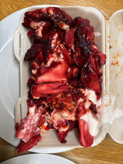 The image shows a styrofoam container filled with char siu pork. The pork is covered in a white sauce. The container is sitting on a wooden surface. The location is Oswestry, United Kingdom.