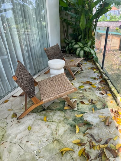 A high-angle view captures a balcony scene in Buduk, Indonesia. Three wooden chairs with woven seats surround a white cylindrical object, possibly a side table or stool. Fallen leaves are scattered on the patterned tile flooring. Lush greenery is visible on the left, contrasting with the urban structures seen through the balcony railing. A glass panel on the left reflects the surroundings, partially obscuring the view. The environment appears tranquil, suggesting a relaxed outdoor setting.