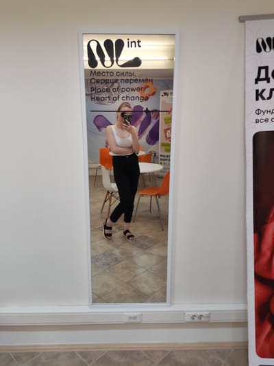 A woman is taking a mirror selfie in a well-lit indoor space, likely a commercial establishment, given the branding visible. She is wearing a white tank top, black pants, and black sandals. Her arms are crossed, and she is holding a smartphone with a patterned case, angled to capture her reflection. The mirror itself is tall and framed in white, reflecting the woman, two orange chairs, and a white table in the background. Behind the mirror, on the wall, a logo with the letters "NL" is displayed prominently, along with text in Russian and English: "Место силы.. Сердце перемен.. Place of power.. Heart of change..". To the right of the mirror, a partial view of a banner with the same "NL" logo is visible. On the wall below the mirror, two electrical outlets are installed in a white conduit. The floor is tiled with a geometric pattern. The scene appears to be located in Nizhnevartovsk, Russia, based on the provided context.