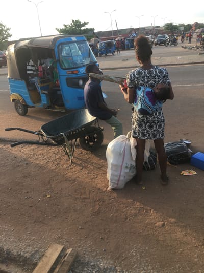 The image shows a daytime scene in Abakaliki, Nigeria, at a roadside location. A woman carrying a child on her back stands next to a blue "keke" (auto-rickshaw). A man sits partly in the keke. Luggage and a large white sack are on the ground near the woman. A wheelbarrow rests on the ground next to the keke. The road is paved but appears rough. Other vehicles and people are visible in the background, and buildings are visible.