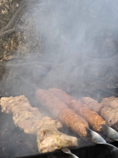 The media file shows a close-up view of meat being grilled over coals, with smoke billowing around it. The scene is dominated by skewers of meat, including two elongated, reddish-brown sausages and pieces of lighter-colored meat, possibly chicken, on other skewers. The meat appears to be seasoned and is in the process of cooking, with some charring and browning visible. The background is a blur of gray smoke and dark, rough textures that suggest a grill or campfire setting. The image captures the sensory experience of cooking meat outdoors. No people are visible, and there is no visible text. The lighting suggests it is daytime. The location context is Pidhorodne, Ukraine, which implies an outdoor setting, likely a barbecue or picnic. The overall impression is one of a rustic, flavorful meal being prepared.
