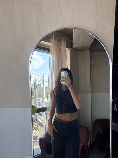 A woman is taking a mirror selfie in a building, likely in Zovuni, Armenia. She is wearing a denim top and jeans with a brown belt. The mirror reflects part of the interior, including chairs and a window with a view of the city. Outside the window are power lines, buildings, and vegetation. The mirror is oval and framed. The wall behind the mirror is light-colored with a textured surface.