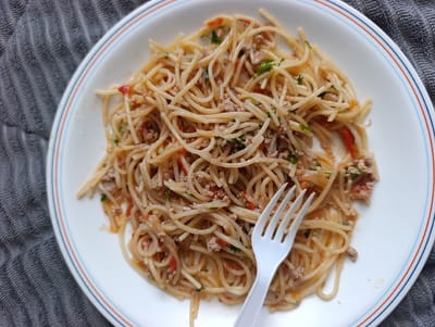 A white, circular ceramic plate with a blue and red rim is positioned on a dark gray, horizontally ribbed textile surface in Kahawa Wendani ward, Kenya. The plate contains a serving of spaghetti coated in a light reddish-brown sauce, mixed with ground meat, diced red vegetables, and green herbs. A white, four-tine plastic fork rests within the spaghetti, angled towards the bottom-left of the plate. No individuals are present in the scene. The lighting is consistent, indicating an indoor setting.