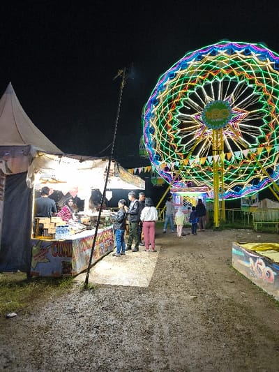 The image displays an outdoor night fair or market scene in An Khe Ward, Vietnam. The ground in the foreground consists of uneven, moist-looking dark earth with minimal sparse grass. On the left, a canvas tent food stall is brightly illuminated, revealing a wooden counter. Behind the counter, one individual in a dark shirt is partially visible, facing away from the camera, and another, wearing a red and white striped top, sits facing forward. The counter displays various items, including water bottles, and a sign prominently featuring "CUMI" in blue letters along with Indonesian text describing "Gorengan Gurita" and pricing. Approximately five adults are gathered in front of this stall.
A tall, slender dark pole with a light fixture at its top stands between the food stall and the background. To the right, a large Ferris wheel is brightly lit with a complex array of green, blue, yellow, and red neon or LED lights, forming a geometric pattern. A circular sign in the center of the Ferris wheel's hub is partially visible, displaying what appears to be "MGO". Several individuals, including adults and possibly children, are observed near the base of the Ferris wheel, some walking or standing. The sky is dark, indicating nighttime, with artificial lighting providing the primary illumination for the scene.