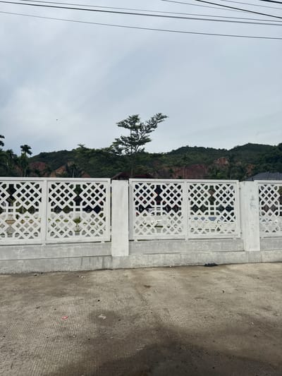 In Lampisang, Indonesia, a white ornamental fence with a geometric lattice pattern runs horizontally across the foreground. Behind the fence, a lush green hillside with scattered trees and rocky outcrops dominates the background. The sky above is overcast and grey, with power lines visible in the upper portion of the frame. The ground in front of the fence is a concrete surface with visible tire tracks and debris. The overall scene depicts a semi-rural or suburban area with natural elements and man-made structures.