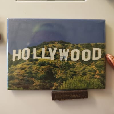 The image shows a rectangular magnet depicting the Hollywood sign on a hillside under a blue and cloudy sky. The magnet is attached to a white surface. A small brown rectangular object is located beneath the magnet. A partially visible, reddish-brown object is on the right side of the magnet. The image does not contain any information relating to the city of Ketoshi, Russia.