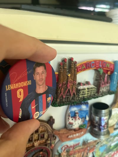 A hand is holding a circular pin that features a portrait of Robert Lewandowski, a soccer player wearing a Barcelona jersey with the number 9. The pin has red and blue stripes, consistent with the Barcelona team colors, and includes his name and signature. To the right of the pin, a collection of miniature replicas of famous landmarks are visible, including the Sagrada Familia, a building with a banner that reads "BARCELONA", and what appears to be a small boat with "ZARAIA" written on it. There is also a metallic mug with some imagery on it, and a plate with "Prague" written on it. The background is out of focus but appears to be a light-colored surface. The scene does not contain any elements or context that would suggest the location is the city of Pokrovnik, Bulgaria.