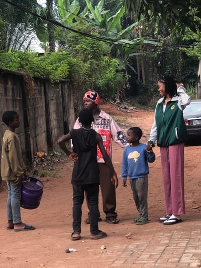 Five individuals stand outdoors on a dirt path next to a building. The first person wears a green and white jacket and pink pants. The second person has on a blue sweatshirt. The third individual has on a pink long-sleeved shirt. The fourth wears black pants and a black shirt. The last individual is carrying a small keg. They appear to be standing at attention. There are trees to the left and a vehicle behind the first person.
