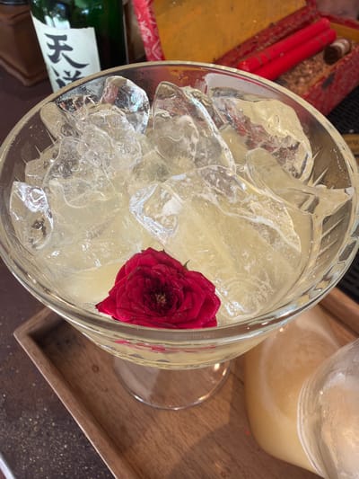 A close-up, overhead shot captures a cocktail filled with ice and a single red rosebud. The drink is served in a clear, stemmed glass set on a wooden tray. In the background, a green bottle with Asian characters and a blurred red box containing red cylindrical objects can be seen. The scene appears to be indoors, possibly a bar or restaurant, located in Baan Thong Krut, Thailand.