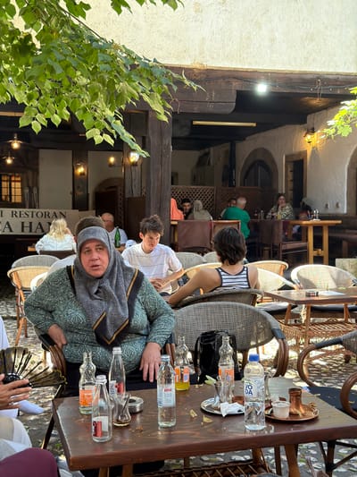 This media file depicts a group of people seated at outdoor tables in a courtyard restaurant. The scene is set in Sarajevo, Bosnia and Herzegovina, with architectural cues like arched doorways and wooden beams suggesting a historical setting. The time of day appears to be daytime, with natural light filtering through the open-air space, supplemented by string lights.
The main subjects are several individuals, some facing the camera, others engaged in conversation or looking away. One woman in the foreground wears a headscarf and a patterned green outfit, looking directly at the camera with a neutral expression. Another person in a striped top is seen from behind. Other individuals are partially visible, seated at various tables.
On the tables, several objects are present, including bottles of water, empty glasses, a copper coffee pot with a small cup, and a black handbag. The overall atmosphere seems relaxed and casual, typical of a cafe or restaurant setting. The ground is paved with small stones.
Visible text on a sign in the background reads "I RESTORAN" and partially "EA HA". The image appears to be taken during a meal or a break at the restaurant. The leaves of a large tree in the foreground add a touch of greenery to the scene.