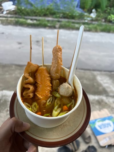 A close-up, slightly elevated perspective captures a person's hand holding a paper cup of Indonesian street food, likely in Wedarijaksa, Indonesia. The white paper cup contains a reddish-brown broth with several items: three skewers, each bearing a different fried item. The leftmost skewer holds a wavy, light brown piece; the middle skewer features a flat, serrated-edged, light brown item; and the rightmost skewer presents an elongated, light brown sausage-like piece. Numerous small, light-colored meatballs and sliced green onions, along with small orange vegetable pieces, are visible within the broth. A white plastic spoon is inserted into the cup. The paper cup rests on a beige ceramic saucer with a dark brown rim, held by a left hand with light to medium skin tone. The background is out of focus, depicting a wet, grey paved surface, possibly a street or sidewalk. Beyond the pavement, green foliage and a blurry blue tarp or plastic sheet are visible. To the right, on the ground, a partially visible discarded cardboard box displays the "AQUA" brand logo, and a pair of dark-colored flip-flops or sandals are present.