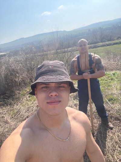 Two adult males are depicted outdoors in a rural setting, identified as Katselovo, Bulgaria. The foreground figure, facing the camera, is a shirtless male with a muscular build, light skin, and short dark hair, wearing a dark bucket hat with a visible logo and a gold-colored chain necklace. He appears to be taking a selfie. Behind him and slightly to the right, a second male with a muscular build, shaved head, and extensive arm tattoos stands holding a two-pronged pitchfork vertically. This second individual is wearing dark blue bib overalls with the straps down, exposing his chest. The ground is covered with dry, yellowish-brown grass and sparse, leafless bushes. In the background, rolling hills with patches of green and brown vegetation extend under a clear blue sky with minimal cloud cover. Both individuals are looking towards the camera.