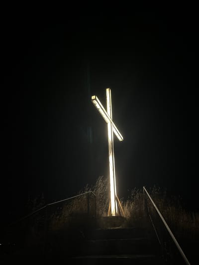 The image depicts a lit cross standing tall against a dark, night sky. The cross is illuminated with a bright, warm light, casting a glow around its structure. The bottom of the cross appears to be anchored on a raised platform, with a set of stairs leading up to it. These stairs are bordered by metal railings on both sides. Tall, dry grass or reeds surround the base of the cross and the stairs, suggesting a natural or somewhat untamed environment. The overall scene is one of solemnity and prominence, with the glowing cross being the central focal point against the deep darkness of the night. There are no people or animals visible in the frame. The location is identified as being in Lozenets, Bulgaria.