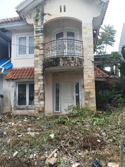The image displays the front façade of a two-story residential building located in Pô Kô, Vietnam, appearing to be in a state of advanced disrepair and neglect. The structure features a cream-colored exterior, heavily stained with dirt, mildew, and water marks, particularly under the upper-level balcony and along the light-colored stone cladding sections.

On the upper floor, an arched balcony with an ornate dark metal railing, featuring scrollwork, extends from the central part of the building. The balcony's concrete floor is discolored and grimy. Behind the railing, white-framed double doors lead into the building. To the left of the balcony, a white-framed window with sheer white curtains is visible. On the ground floor, a white wooden front door with a vertical panel design is flanked by narrow vertical windows. To the left of the main entrance, a second ground-floor window, also white-framed, shows signs of broken or missing glass and has a metal grate visible behind it; above this window is a small overhang with terra cotta roof tiles. A portion of an adjacent structure with a blue corrugated roof and a white gutter is visible on the far left. The roofline and upper walls of the depicted building exhibit some vegetation growth.

The foreground consists of a heavily neglected and overgrown area. The ground is covered with dry weeds, brush, and a substantial accumulation of debris including small construction fragments, plastic refuse, and discarded organic matter such as cut branches and leaves. No individuals, actions, or interactions are visible in the scene.