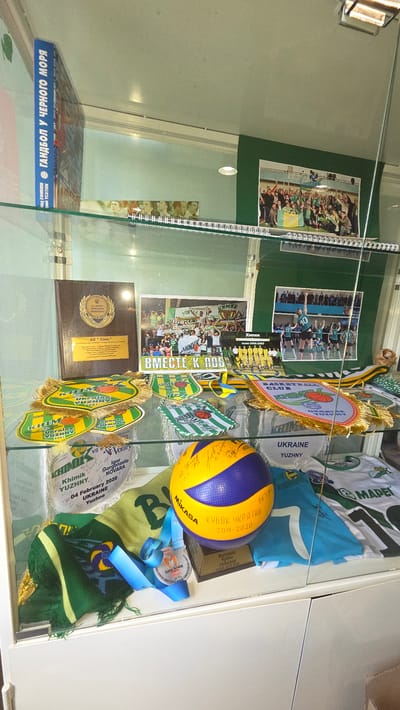 This media file captures a display case containing memorabilia related to a Ukrainian volleyball club, likely the "Khimik" club from Yuzhny. The display is oriented vertically and features various objects, including team photographs, banners, medals, and a volleyball.
The subjects are primarily objects associated with the club's history. Several team photos are visible, showing groups of athletes in uniform, some celebrating victories. Banners with the club's name and affiliations like "UKRAINE YUZHNY" are prominently displayed. A yellow and blue volleyball, signed by various individuals, is a central object. Medals with blue ribbons, some indicating achievements like "КУБОК УКРАЇНИ 2019-2020" (Ukrainian Cup 2019-2020), further highlight the team's accomplishments. Other items include scarves and pennants with team logos and slogans.
The overall impression is that of a sports history exhibit, likely in a museum, arena, or club facility. The setting appears to be indoors, with glass casing protecting the items. There are no clear indicators of the time of day or weather, but the lighting suggests an indoor environment. The emotions conveyed are those of pride and achievement, as evidenced by the celebratory photos and award displays. The visible text includes team names, years, competition titles, and locations within Ukraine. The book at the bottom left appears to be about "Volleyball on the Black Sea" (ГАНДБОЛ У ЧЕРНОГО МОРЯ).