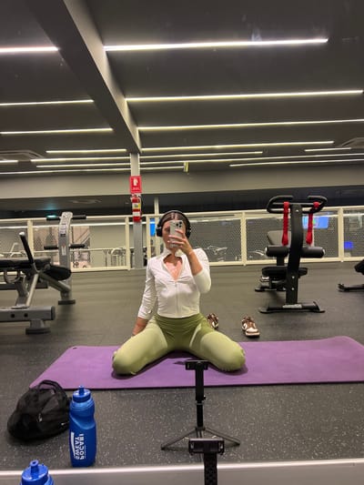 The image depicts a woman with dark hair and headphones taking a selfie in a gym. She is wearing a white zip-up long-sleeve shirt and olive green leggings. She is kneeling on a purple yoga mat, with a black gym bag and a blue water bottle to her left. Her shoes, which appear to be brown and white sneakers, are visible to her right. The gym environment features various weight machines and equipment, with a grey ceiling and modern strip lighting. A fire extinguisher is mounted on a pillar in the background. The scene is set in a gym in Si Sunthon, Thailand.