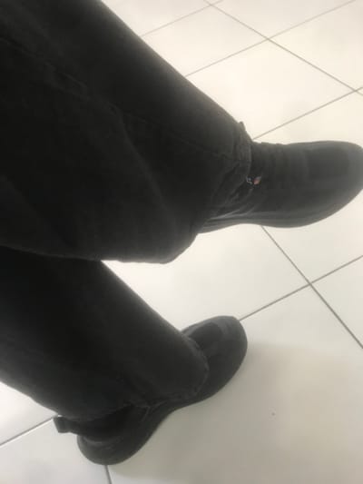 An overhead view captures the lower legs and feet of an individual, seated or standing. The person wears dark, likely black, trousers that are visibly creased, and dark, presumably black, athletic shoes with thick soles. A small, rectangular tag with red, white, and blue elements is visible near the cuff of the right trouser leg, above the shoe. The legs are bent, and the right leg appears to be positioned slightly in front of the left. The footwear rests on a light-colored, reflective floor composed of large, square white tiles with dark grout lines, forming a grid pattern. The environment is uniformly lit, indicating an indoor setting. The specific city of Muka, Indonesia is not visually discernible within the image.