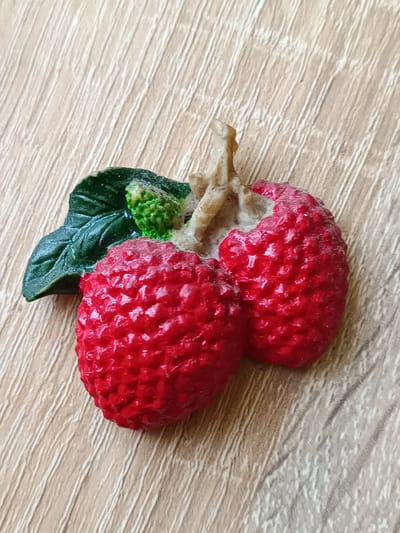 A decorative figurine of two red lychees with green leaves and a stem rests on a light brown wooden surface. The lychees have a textured, bumpy surface and are split open, revealing a white interior. The leaves are dark green and detailed, and the stem is light brown and twig-like. The background surface appears to be a laminate or wood with visible grain lines running diagonally. The scene is lit from above and to the right, casting subtle shadows. There are no people or any other objects present in the image. The image is a close-up shot, focusing entirely on the lychee figurine and the surface it rests on. This scene does not have any relation to Tebras, Latvia.