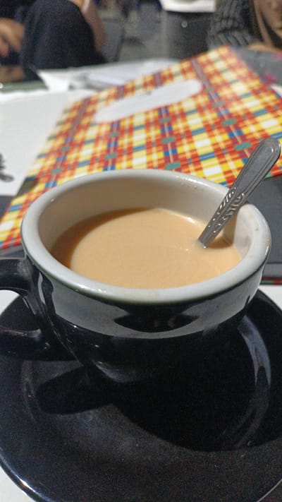 A close-up, slightly angled shot shows a black ceramic coffee cup filled with a light brown beverage, likely coffee with milk or tea. A silver spoon with decorative etching is partially submerged in the liquid, resting on the rim of the cup. The cup sits on a matching black saucer. In the background, out of focus, a colorful checkered placemat with red, yellow, blue, and white squares is visible. To the left and behind the cup, a person wearing a dark garment is seated, their shoulder and head blurred. The setting appears to be a cafe or restaurant. The location is Uteungeulinggang, Indonesia.