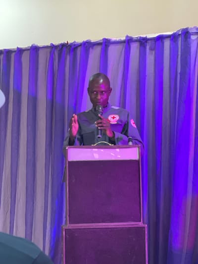 The image shows a man in a dark uniform with red cross insignia on the sleeves and chest, standing behind a podium. He appears to be speaking into a microphone. The man is facing towards the right of the frame, with his left hand near the microphone and his right hand gesturing. The background consists of horizontal purple and white striped fabric drapes, illuminated by purple lighting, creating a stage-like setting. Two large black speakers are positioned behind the podium to the right. The overall scene suggests a formal event or presentation. The man's expression is difficult to discern due to the image quality and angle, but he seems engaged in his speech. The lighting and drapes suggest an indoor event, possibly in the evening. There is no visible text in the image, besides the red cross symbol. The location context suggests the event is taking place in Unguwar Sarki, Nigeria.