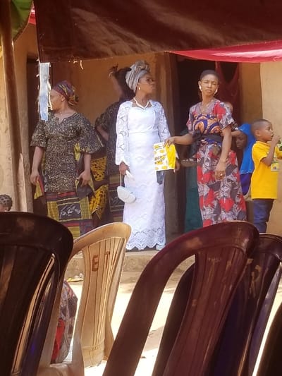 The image captures an outdoor social gathering in Ochadam, Nigeria. The scene is set in front of a light-brown building with an open doorway, above which dark brown and red fabrics are suspended, creating a makeshift canopy.
In the midground, five individuals are centrally positioned. From left, a woman with dark skin wears a patterned headscarf (brown, red, yellow) and a black, gold, and red patterned dress, looking left. Next to her, a woman with dark skin wears a light grey headwrap, a white long-sleeved lace dress, and a pearl necklace. She holds a small white handbag in her left hand and a yellow plastic bag labeled "Goodies" in her right, looking upwards and to the right. To her right, a third woman with dark skin and short dark hair wears a red, blue, and black floral patterned dress with a black waist sash, facing forward. To the far right, a young boy in a yellow shirt and blue trousers stands, holding an object to his mouth. A smaller child, in blue, is partially visible behind the third woman. Additional individuals are partially visible in the blurred background within the building's doorway.
The foreground is occupied by three plastic chairs: a dark brown/black chair on the left, a light brown chair with "ETU" inscribed on its backrest in the center, and a dark brown/red chair on the right, partially obscuring the lower bodies of the subjects. The ground is composed of light brown packed earth. The lighting indicates bright daylight.