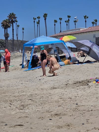 The image depicts a sunny day at a beach in San Clemente, California, United States of America. Several people are present, some lounging under beach umbrellas and canopies, others standing. In the foreground, two women are interacting. One woman in a striped bikini is crouching down, appearing to hold or tend to a younger child, also in a bikini. Beside them, a woman in a black swimsuit and a green baseball cap stands facing away from the camera.

Further back, a man in swim trunks is seated under a large gray umbrella. To the left, a person in a bright orange hooded jacket and swim trunks stands holding a phone. The beach is sandy with scattered small stones. Palm trees are visible in the background, along with a building with a tiled roof and a security camera. Beach equipment, including umbrellas and a blue and white striped canopy, are set up on the sand. A few colorful toy building blocks are visible in the lower right corner. The sky is clear and blue.