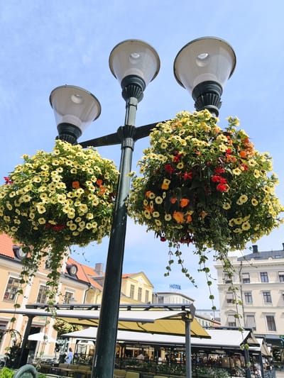 The image captures a street scene in Linköping, Sweden, featuring a prominent lamppost adorned with two large hanging baskets of yellow and orange flowers. The lamppost itself is dark green with ornate detailing on the light fixtures and pole. In the background, several buildings are visible: a yellow building with red-tiled roofs to the left, and a lighter-colored building with multiple windows to the right, which appears to be a hotel as indicated by a blue "HOTEL" sign. Below the lamppost and extending into the foreground is an outdoor seating area for a cafe or restaurant, complete with tables, chairs, and umbrellas, some of which are yellow. Several people are seated at the tables, and a few individuals are walking in the background. The sky is a clear blue with sparse clouds. The overall impression is a bright, sunny day in a European city.
