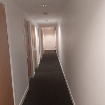 The image depicts a dimly lit, neutral-toned corridor within an unidentified building. The walls are painted a light shade of white or cream, contrasting with the dark gray or black carpet covering the floor. Multiple doors, some partially visible, line the left side of the corridor, suggesting individual rooms or units. Several ceiling-mounted light fixtures are present, though their contribution to the overall illumination appears minimal. The perspective of the photo implies a vanishing point towards the end of the corridor, with the lighting becoming progressively brighter in that direction. No people or specific objects are discernible within the frame. The location, though stated as Coventry, United Kingdom, is not identifiable from the image alone; there are no specific features visible that would confirm the building's whereabouts within the city.