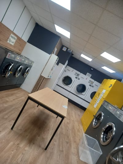 The image depicts a laundromat with several rows of washing machines. Some are yellow and gray, while others are dark gray. A small table with a light brown top and black legs is positioned on the wooden floor, near a transparent plastic container. The overall impression is a clean, commercial laundry facility.