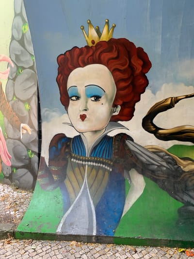 This media file displays a mural painted on an outdoor surface, likely a pavement or wall, given the adjacent cobblestone path. The mural features a stylized, almost doll-like portrait of a figure with striking blue eyeshadow and red lips. This figure wears a golden crown and has voluminous, curly red hair. Parts of a winged creature, possibly a bird or a fantastical being, are visible behind the figure, rendered in browns and grays. The background transitions from a soft blue sky with white clouds to a vibrant green landscape. The lower portion of the mural depicts a scene with grey rocks and hints of pink organic shapes, suggesting a fantastical or mythological setting. The artwork appears to be situated in an urban environment, as evidenced by the cobblestones and the overall context of public art. The lighting suggests daylight, but the time of day is not precisely determinable. The artwork's aesthetic is bold and graphic, with a surreal quality. No visible text is present within the mural itself. The location context of Odivelas, Portugal, suggests this is a public art installation within that municipality.