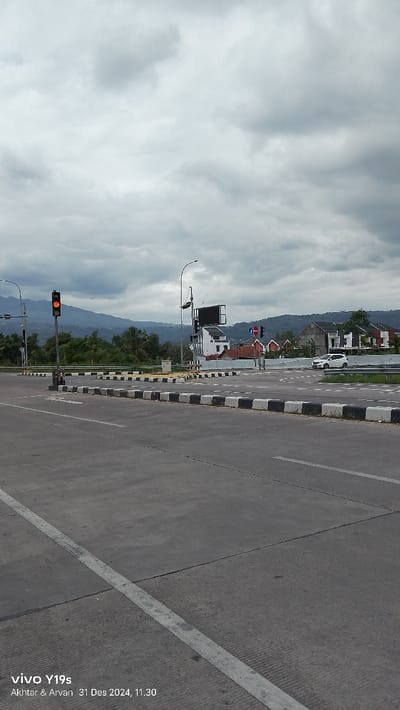 The image presents a wide outdoor view of a modern road intersection or interchange, likely in Sumedang, Indonesia, on an overcast day. In the foreground, a multi-lane concrete road stretches forward, marked with white lines defining lanes and an approaching intersection. The median strips and traffic islands within the intersection are clearly delineated by black and white painted curbs.
A prominent traffic light pole on the left displays an illuminated amber (yellow) light, indicating a cautionary state. Further into the intersection, other traffic signals and a blue circular "no entry" sign are visible. To the right of the intersection, a white car is seen, possibly navigating or waiting. On the right side, beyond the road, several modern, light-colored buildings with dark roofs are nestled amidst green foliage. In the mid-ground, a large, dark rectangular sign or billboard is mounted on a pole.
The backdrop consists of a range of distant, green-hued mountains under an entirely overcast sky filled with diffused grey clouds, suggesting a muted daylight. No people are visible, lending the scene a quiet, somewhat empty atmosphere. The bottom left corner of the image contains a watermark: "vivo Y19s Akhtar & Arvan 31 Des 2024, 11.30", providing details about the device, photographers, and the time the photograph was taken.