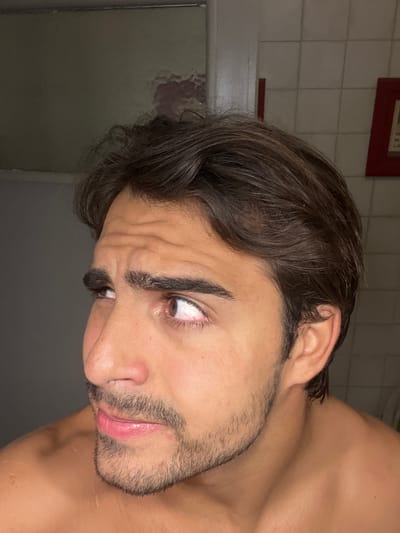 A close-up, sideways portrait features a young man from the chest up, shirtless. His head is sharply tilted, with his left eye (viewer's right) wide open and looking intensely upwards, revealing a significant amount of white sclera below the iris. His eyebrows are deeply furrowed, causing prominent wrinkles across his forehead, indicating a strained or focused expression. He has dark, short hair and a noticeable stubble or short beard. His lips are slightly parted. The background consists of a light-colored tiled wall with dark grout lines, suggesting an indoor setting. A dark red rectangular object, possibly a small frame, is visible on this wall behind his head. To the left, the background is darker and indistinct. The lighting is artificial, highlighting his features. No discernible text is visible.