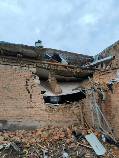 The image displays the severely damaged exterior of a brick building located in Pysarivka, Ukraine, under an overcast sky. The structure exhibits extensive destruction, with a large, irregular breach in its upper brick wall, exposing interior elements. Below this opening, a significant accumulation of rubble, primarily composed of broken red bricks, splintered wooden debris, and twisted metal fragments, lies at the base of the wall. The roof directly above the breach is compromised, showing shattered framing, torn roofing materials, and a large, bent metal pipe. A detached, L-shaped concrete or plaster section hangs precariously from the remaining ceiling structure within the exposed interior. To the right, a bent and twisted metal support frame rests among the debris. No individuals are present in the scene.