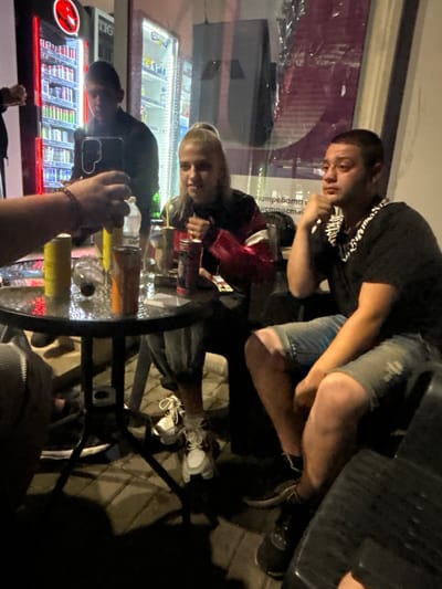A group of people are gathered around a round glass-top table at night. In the background, illuminated refrigerated display cases are visible, suggesting a retail or convenience store setting, likely within the city of Krivodol, Bulgaria, given the context.

On the left, a person's arm and hand, wearing a bracelet, are visible reaching towards the table, holding a bottle. To the right of this arm, a younger individual with blonde hair tied in a ponytail is seated, wearing a black top with a red pattern. They are looking towards the right side of the frame.

In the center of the frame, partially obscured by the table, is a person with short dark hair wearing a dark jacket. They appear to be looking down.

To the right of the table, a man with short dark hair is seated, wearing a black t-shirt with a distinctive striped collar accessory and denim shorts. He is leaning forward, with his chin resting on his hand, looking towards the right.

The table itself is cluttered with various items, including cans (one yellow, one orange, and one red), a partially empty plastic bottle, and what appears to be a napkin. The ground appears to be paved with interlocking bricks. The lighting suggests artificial illumination from the store's displays and possibly ambient streetlights. The overall atmosphere seems casual, possibly a break or stop at a store during an evening outing.